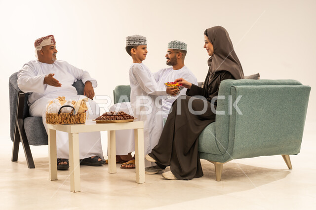 زينة وديكورات الأعياد ، أجواء العيد في المجتمع العماني ، مشاركة اجواء رائعة لعائلة عمانية داخل المنزل تحتفل بالعيد ، الزيارات العائلية وتجمعات الأسر ، ابن يقدم الضيافة والحلوى لوالدته ، بورتريه لعائلة عربية خليجية عمانية تحتفل بالأعياد , خلفية بيضاء .