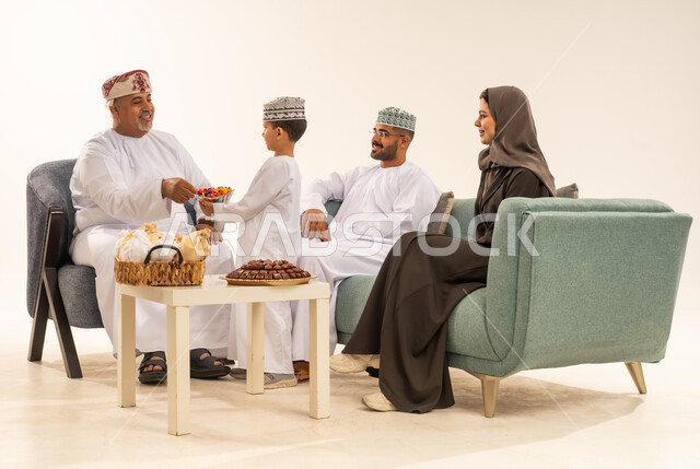 أعياد ومناسبات إسلامية سعيدة, تبادل المعايدات بين العائلة ، مشاركة اجواء رائعة لعائلة عمانية داخل المنزل تحتفل بعيد الفطر السعيد ، الزيارات العائلية وتجمعات الأسر ، تقديم الضيافة و الحلوى ، بورتريه لعائلة عربية خليجية عمانية تحتفل بالأعياد , خلفية بيضاء .