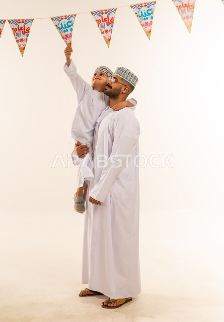 Celebration rituals and happy occasions, the tenderness of fatherhood and a close-knit family, feelings of affection and family love. Portrait of an Arab Gulf Omani family celebrating Islamic holidays. A father carrying his son, both wearing a dishdasha and a kummah, hanging decorative strings. Full-length body, white background....