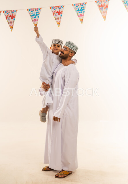 ديكورات وزينة رمضانبة ، ملاعبة الأبناء وملاطفتهم , بناء علاقة قوية مع الأولاد، بورتريه لعائلة عربية خليجية عمانية تحتفل بالأعياد الإسلامية, أب يحمل ابنه يرتديان الدشداشة والكمه يقومان بتعليق حبال الزينة، قضاء اوقات ممتعه ، الجسم بالطول الكامل، خلفية بيضاء .