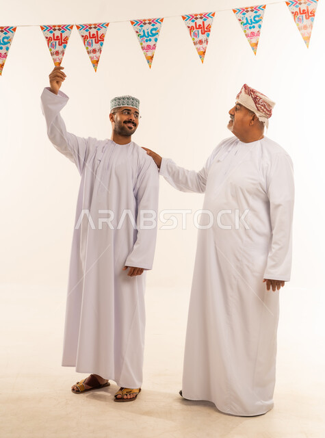 مشاعر المودة والمحبة العائلية, طقوس الاحتفالات والمناسبات السعيدة في سلطنة عمان، بورتريه لعائلة عربية خليجية عمانية تحتفل بالأعياد الإسلامية, أب و ابنه يرتديان الدشداشة والكمه يقومان بتعليق حبال الزينة، الجسم بالطول الكامل، خلفية بيضاء .