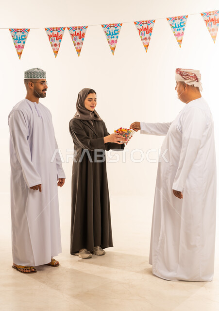 الاحتفال بقدوم العيد ، تقديم الضيافة وحلوى العيد , ديكورات وزينة رمضانية ، إظهار مشاعر المودة والمحبة العائلية, بورتريه لعائلة عربية خليجية عمانية تحتفل بالأعياد الاسلامية, امراة عربية خليجية عمانية تقدم الضيافة فى العيد ، صورة الجسم بالطول الكامل ، خلفية بيضاء.