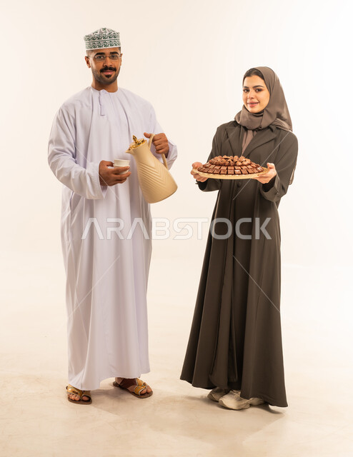 النظر الى الكاميرا بايماءات الفرحة والسرور والسعادة بالاحتفاليات ، بهجة وفرحة العيد, حسن استقبال واكرام الضيوف, العيد بين الاهل , مناسبات واعياد اسلاميه, تقديم الضيافة العربية ، زيارات وتجمعات عائليه, زوجان عربيان عمانيان خليجيان يقدمان الضيافة ، صورة الجسم بالطول الكامل ، خلفية بيضاء .