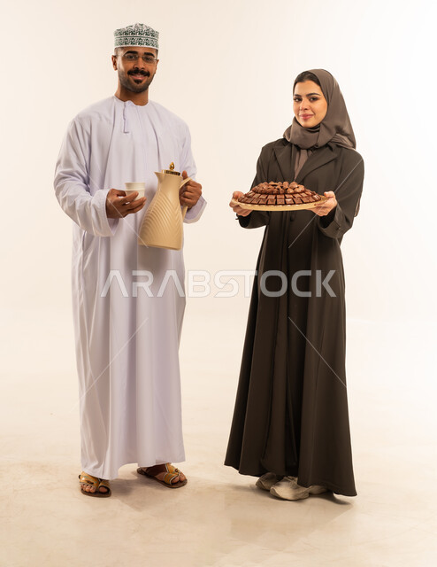 المعايدات والتهنئات وطقوس المناسبات الاسلامية، النظر الى الكاميرا بايماءات الفرحة والسرور والسعادة بالاحتفاليات ، بهجة وفرحة العيد, حسن استقبال واكرام الضيوف, العيد بين الاهل , تقديم الضيافة العربية ، زيارات وتجمعات عائليه, زوجان عربيان عمانيان خليجيان يقدمان الضيافة ، صورة الجسم بالطول الكامل ، خلفية بيضاء .