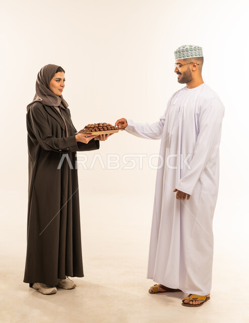 تعبيرات الفرحة و السرور و السعادة بالاحتفاليات, حسن الضيافة في المناسبات و الأعياد الاسلامية ، امراه عربيه عمانية خليجيه تقوم بتقديم حلوى العيد لزوجها ، الاحتفال بقدوم العيد ، صورة الجسم بالطول الكامل ،خلفية بيضاء .