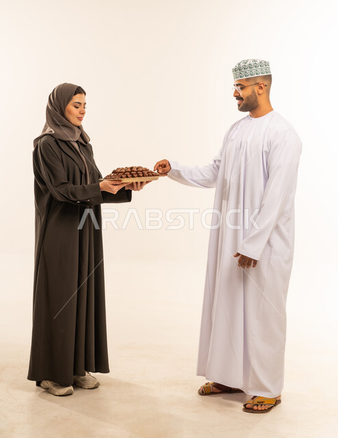 اكرام الضيف وحسن الاستقبال ، مشاركة العائلة الحلوى و المأكولات الشهية ، مناسبات واعياد اسلاميه, زيارات وتجمعات عائليه, تعبيرات الفرحة و السرور و السعادة بالاحتفاليات,  امراه عربيه عمانية خليجيه تقوم بتقديم حلوى العيد لزوجها ، الاحتفال بقدوم العيد ، صورة الجسم بالطول الكامل ،خلفية بيضاء .