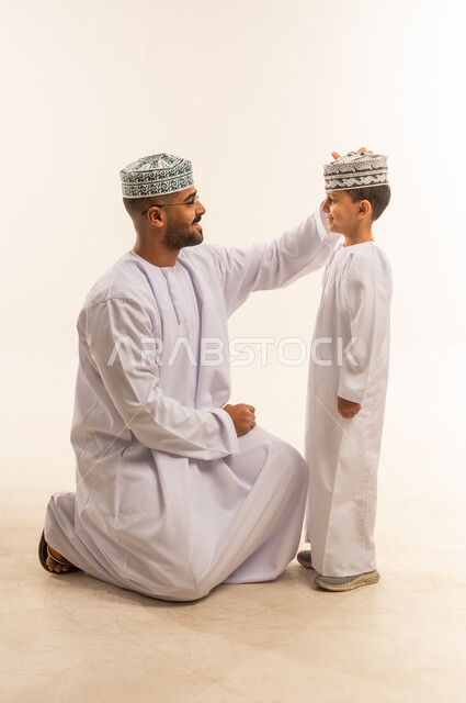 قضاء اوقات ممتعه مع الابناء ، أجواء أسرية مليئة بالمودة، حنان الابوة والاسرة المتماسكة ، بناء علاقة قوية مع الأولاد، ملاعبة الأبناء وملاطفتهم , بورتريه لرجل عربي خليجي عماني يرتدي الدشداشة والكمة  يجلس على ركبتيه ويلاعب ابنه ، الجسم بالطول الكامل، خلفية بيضاء .
