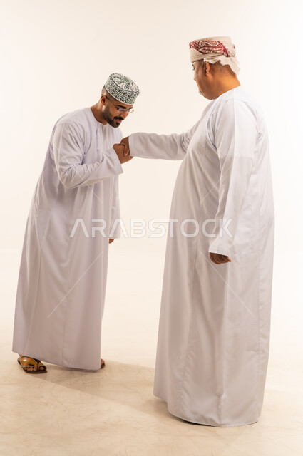 The concept of honoring and appreciating parents, feelings of love, affection and fatherhood, a portrait of an Arab Gulf Omani man wearing a dishdasha and a kuma greeting his elderly father on Eid, kissing the father's hand and congratulating him on the advent of Eid, wearing traditional Omani clothing on Islamic holidays, white background.