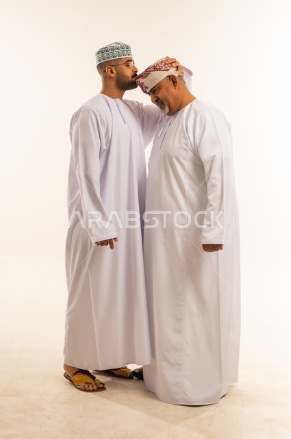 تقبيل رأس الأب والتهنئة بقدوم العيد، بورتريه لرجل عربي خليجي عماني يرتدي الدشداشة والكمه يقوم بمعايدة والده كبير السن في العيد، مشاعر الحب والمودة والأبوة، مفهوم بر الوالدين وتقديرهم، ارتداء الملابس العمانية التقليدية في الأعياد الإسلامية، خلفية بيضاء .