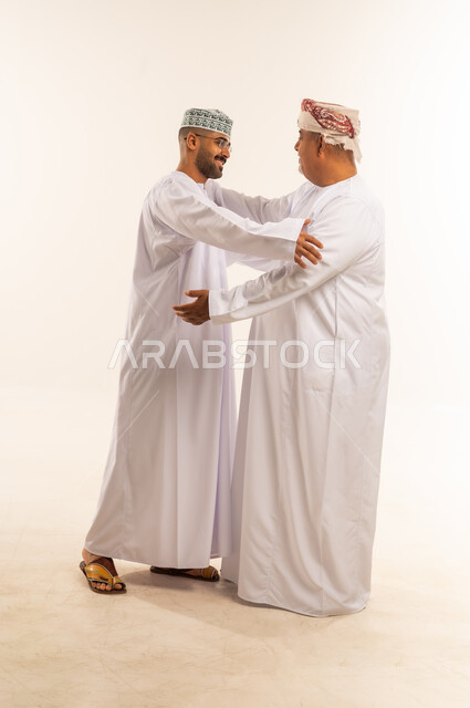 تقبيل الأب والتهنئة بقدوم العيد، الاحترام والتقدير للوالدين ورد الجميل لهما، مشاعر الحب والمودة والأبوة، بورتريه لرجل عربي خليجي عماني يرتدي الدشداشة والكمه يقوم بمعايدة والده كبير السن في العيد، يقوم باحتضان ابنه بحب وحنان ، ارتداء الملابس العمانية التقليدية في الأعياد الإسلامية، خلفية بيضاء .