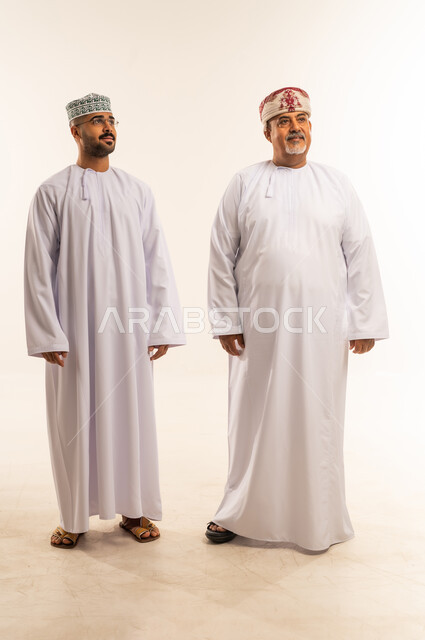 Looking at something with gestures of happiness, a father's great love for his children, paternal bond and family cohesion, gestures of love and affection for children, portrait of an Arab Gulf Omani father and son wearing the dishdasha and the kummah, wearing traditional Omani clothing on Islamic holidays, white background.