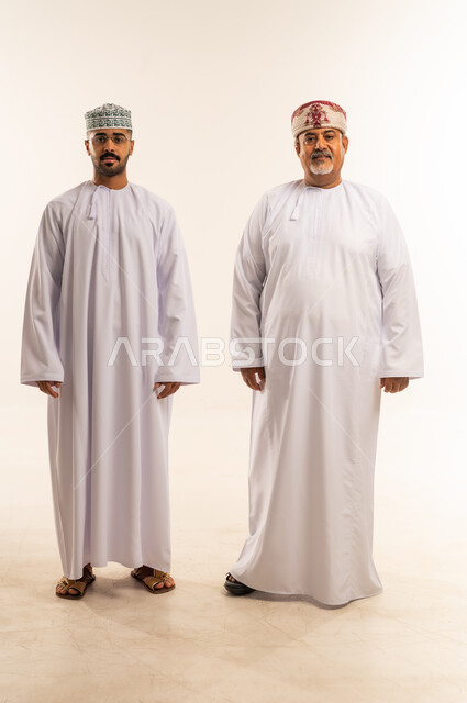 ارتداء الملابس العمانية التقليدية في الأعياد الإسلامية، النظر الى الكاميرا بايماءات السعادة ، حب الأب الكبير لأولاده , الرابطة الأبوية والترابط الأسري، ايماءات الحب والعطف على الأبناء، بورتريه لأب وابنه عربيان خليجيان عمانيان يرتديان الدشداشة والكمة ، خلفية بيضاء .