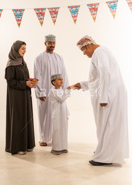 أجواء البهجة والسعادة والاحتفال, إظهار مشاعر المودة والمحبة العائلية, السلام وتقبيل رأس الجد والتهنئة بقدوم العيد, ديكورات وزينة رمضانية ، بورتريه لعائلة عربية خليجية عمانية تحتفل بالأعياد الاسلامية وتتبادل المعايدات, تقديم حلوى العيد ، خلفية بيضاء.