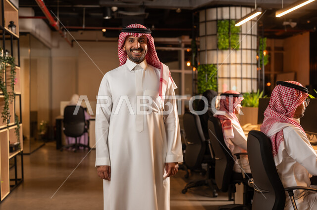 النظر الى الكاميرا بايماءات الثقة بالنفس، العمل في شركات المملكة العربية السعودية، مفهوم الاناقة والرجولة والاهتمام بالمظهر الخارجي، رجل أعمال عربي خليجي سعودي مبتسم يرتدي الشماغ والثوب التقليدي يقف باستقامة داخل المكتب