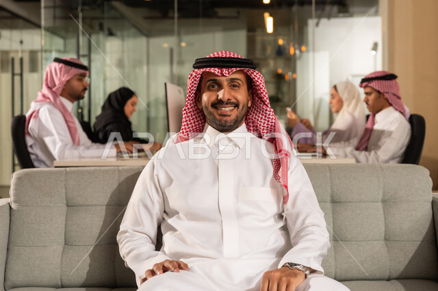 Working in a Saudi Arabian company, looking at the camera with gestures of happiness and self-confidence, the concept of elegance, masculinity and interest in external appearance, a smiling Saudi Arabian Gulf businessman wearing a traditional shemagh and thobe sitting on a comfortable gray sofa inside the office, a work team meeting