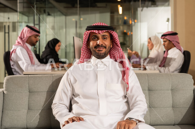 اجتماع فريق العمل،  شركات المملكة العربية السعودية، النظر الى الكاميرا بايماءات السعادة الثقة بالنفس، مفهوم الاناقة والرجولة والاهتمام بالمظهر الخارجي، رجل أعمال عربي خليجي سعودي مبتسم يرتدي الشماغ والثوب التقليدي يجلس على كنبة رمادية مريحة داخل المكتب