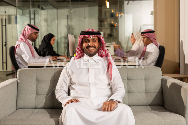 العمل في شركات المملكة العربية السعودية، النظر الى الكاميرا بايماءات السعادة الثقة بالنفس، مفهوم الاناقة والرجولة والاهتمام بالمظهر الخارجي، رجل أعمال عربي خليجي سعودي مبتسم يرتدي الشماغ والثوب التقليدي يجلس على كنبة رمادية مريحة داخل المكتب، اجتماع فريق العمل