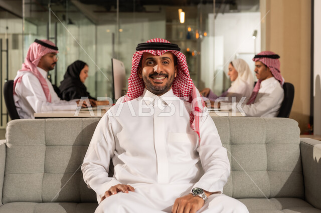 اجتماع فريق العمل،  شركات المملكة العربية السعودية، النظر الى الكاميرا بايماءات السعادة الثقة بالنفس، مفهوم الاناقة والرجولة والاهتمام بالمظهر الخارجي، رجل أعمال عربي خليجي سعودي مبتسم يرتدي الشماغ والثوب التقليدي يجلس على كنبة رمادية مريحة داخل المكتب