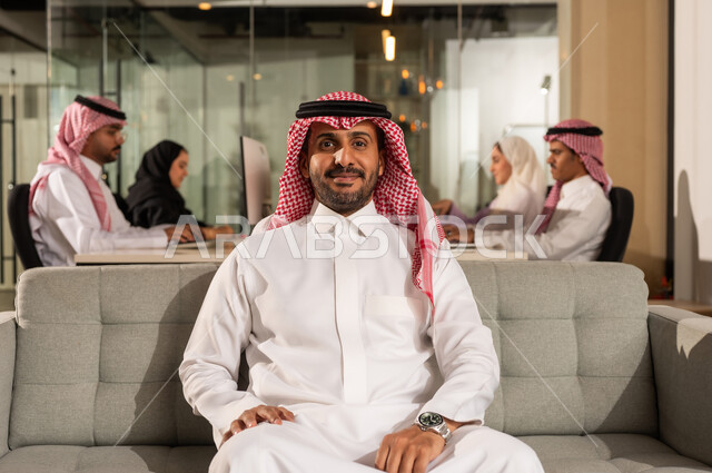 العمل في شركات المملكة العربية السعودية، النظر الى الكاميرا بايماءات السعادة الثقة بالنفس، مفهوم الاناقة والرجولة والاهتمام بالمظهر الخارجي، رجل أعمال عربي خليجي سعودي مبتسم يرتدي الشماغ والثوب التقليدي يجلس على كنبة رمادية مريحة داخل المكتب، اجتماع فريق العمل