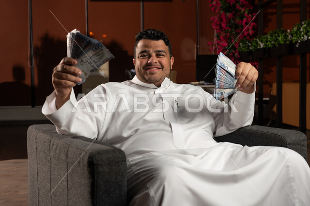 مفهوم الوفرة المالية، التخطيط في تمويل المشاريع المستقبلية, رجل أعمال عربي خليجي سعودي مبتسم يرتدي الثوب التقليدي يجلس على كنبة مريحة بإيماءات السرور والسعادة بالإدخار المالي, النمو والأرباح المالية, الاستثمار في الاقتصاد السعودي