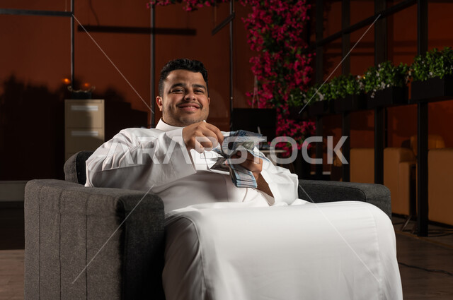 مفهوم الوفرة المالية، التخطيط في تمويل المشاريع المستقبلية, رجل أعمال عربي خليجي سعودي مبتسم يرتدي الثوب التقليدي يجلس على كنبة مريحة بإيماءات السرور والسعادة بالإدخار المالي, النمو والأرباح المالية, الاستثمار في الاقتصاد السعودي