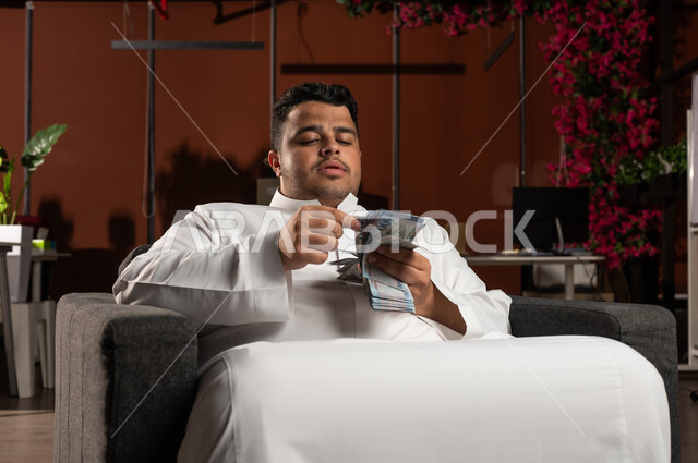 مفهوم الوفرة المالية، التخطيط في تمويل المشاريع المستقبلية, رجل أعمال عربي خليجي سعودي مبتسم يرتدي الثوب التقليدي يجلس على كنبة مريحة بإيماءات السرور والسعادة بالإدخار المالي, النمو والأرباح المالية, الاستثمار في الاقتصاد السعودي