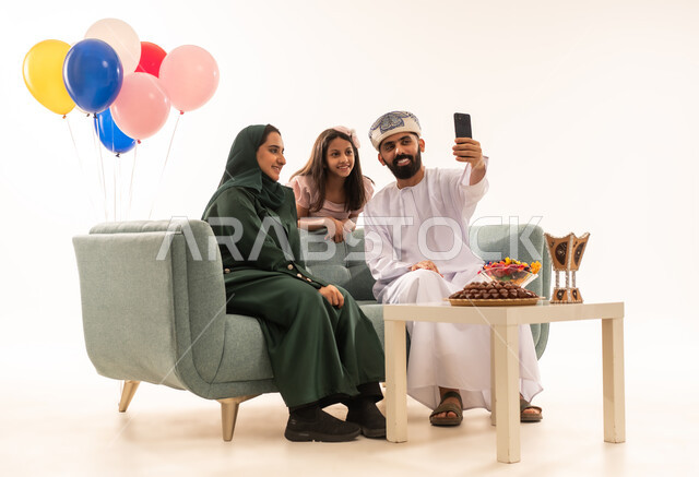 التقاط صور السيلفي التذكاريه ، الزيارات العائلية وتبادل التهنئات والمعايدات بين أفراد الأسرة بإيماءات الفرح والسرور, بورتريه لعائلة عربية خليجية عمانية تحتفل بالعيد السعيد, تقديم الحلويات والضيافة الشعبية، طقوس الأعياد والمناسبات الاسلامية في سلطنة عمان ، خلفية بيضاء .