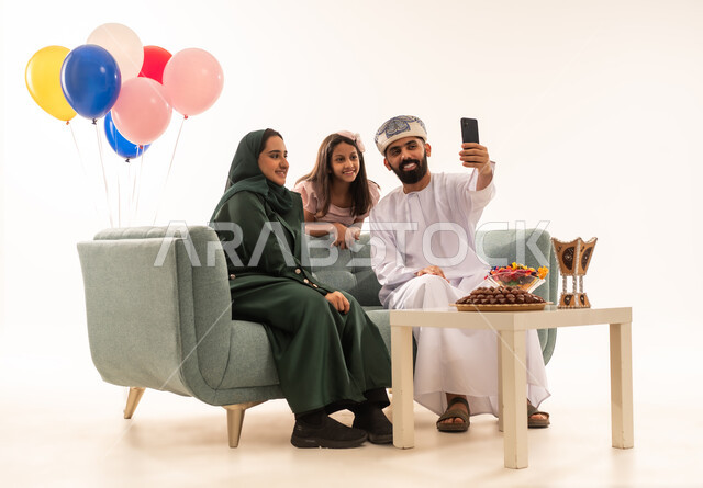 توثيق اللحطات التذكارية ، تقديم الحلويات والضيافة الشعبية، الزيارات العائلية وتبادل التهنئات والمعايدات بين أفراد الأسرة بإيماءات الفرح والسرور, بورتريه لعائلة عربية خليجية عمانية تحتفل بالعيد السعيد, طقوس الأعياد والمناسبات الاسلامية في سلطنة عمان ، خلفية بيضاء .