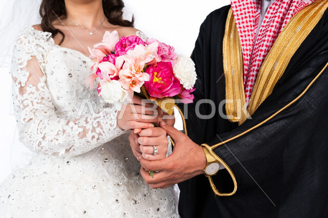 بورتريه بصورة مقربة لعريس بالزي السعودي الرسمي يمسك يد عروسته ، عروسة عربية خليجية سعودية جميلة ، ترتدي بدلة الزفاف البيضاء ، تحمل بيدها باقة ورد جميلة ، جلسة تصوير رائعة لعريسان سعوديان في يوم زفافهما ، ليلة العمر، مناسبات وأفراح