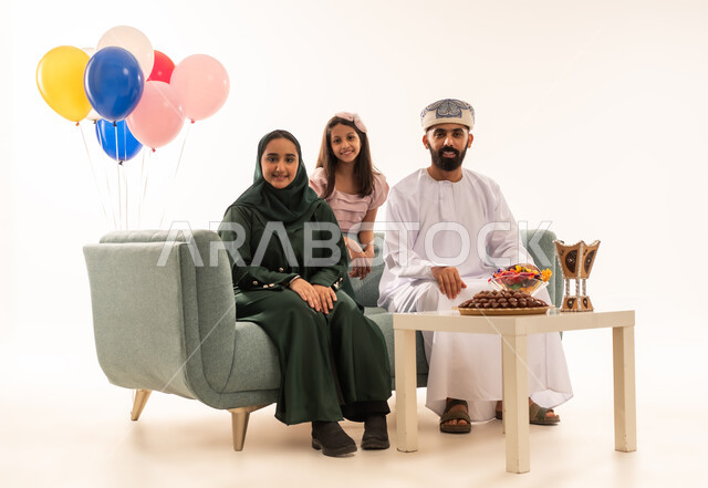 طقوس الأعياد والمناسبات الاسلامية في سلطنة عمان ، الجلوس في غرفة المعيشة في جو عائلي دافئ، بورتريه لعائلة عربية خليجية عمانية تحتفل بالعيد السعيد, الزيارات العائلية وتبادل التهنئات والمعايدات بين أفراد الأسرة بإيماءات الفرح والسرور، تقديم الحلويات والضيافة الشعبية, خلفية بيضاء .