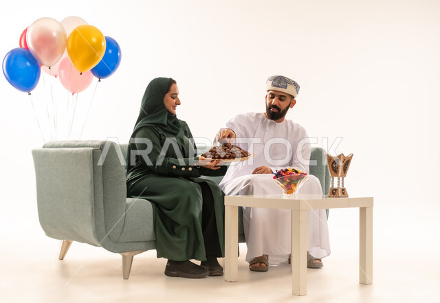 مفهوم الحنان والمحبة والمودة، تقديم الضيافة العربية والحلويات الشعبية، الزيارات العائلية والاحتفال بالأعياد الإسلامية، بورتريه لعائلة عربية خليجية عمانية تجلس في غرفة المعيشة ، امرأة عربية عمانية تقوم بتقديم التمر لزوجها , إظهار مشاعر الفرح والسعادة بقدوم العيد، خلفية بيضاء .