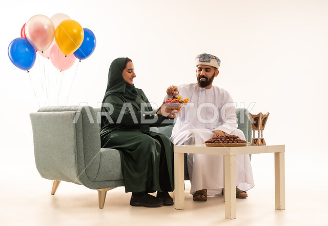 امرأة عربية عمانية تقوم بتقديم حلوى العيد لزوجها , اجواء عائلية دافئة ، تقديم الضيافة العربية والحلويات الشعبية، الزيارات العائلية والاحتفال بالأعياد الإسلامية، مفهوم الحنان والمحبة والمودة،  بورتريه لعائلة عربية خليجية عمانية تجلس في غرفة المعيشة ، إظهار مشاعر الفرح والسعادة بقدوم العيد، خلفية بيضاء .