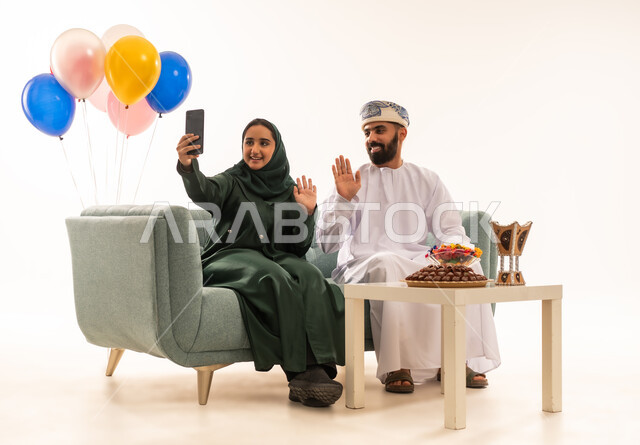 استخدام الهاتف المحمول، التقاط صور سيلفي تذكارية، زوجان عمانيان عربيان في غرفة المعيشة، اجواء العائلة العمانية في العيد، اللمة العائلية، العيد بين الاهل في المجتمع العماني ، زيارات العيد، اعياد ومناسبات ، خلفية بيضاء .