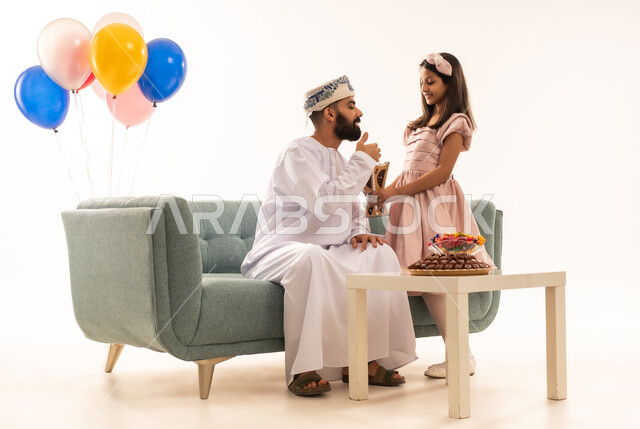 التبخير واستخدام عطور العود الفاخرة ، تقديم العيدية للأطفال في عيد الفطر المبارك ، الزيارات العائلية وتجمعات الأسر ، تقديم العيديات و الحلوى ،  جو البهجة والفرح والزينة، بورتريه لعائلة عمانية داخل المنزل تحتفل بعيد الفطر السعيد ، عيد فطر مبارك ، خلفية بيضاء .