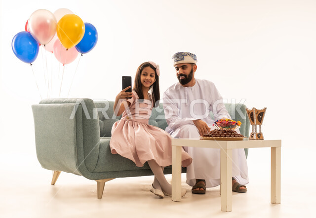 استخدام الجوال فى توثيق اللحظات التذكارية فى العيد ، تقديم العيدية للأطفال في عيد الفطر المبارك ، الزيارات العائلية وتجمعات الأسر ، تقديم العيديات و الحلوى ،  جو البهجة والفرح والزينة، بورتريه لعائلة عمانية داخل المنزل تحتفل بعيد الفطر السعيد ، عيد فطر مبارك ، خلفية بيضاء .