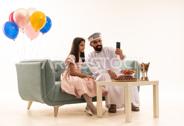استخدام التكنولوجية والتقنيات الحديثة ، تقديم العيدية للأطفال في عيد الفطر المبارك ، الزيارات العائلية وتجمعات الأسر ، تقديم العيديات و الحلوى ،  جو البهجة والفرح والزينة، بورتريه لعائلة عمانية داخل المنزل تحتفل بعيد الفطر السعيد ، عيد فطر مبارك ، خلفية بيضاء .