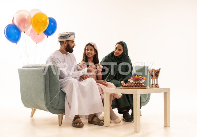 المودة والمحبة العائلية ، مفهوم الكرم وتقديم الضيافة العربية، طقوس الاحتفالات والمناسبات السعيدة في سلطنة عمان، جو البهجة والفرح والزينة، بورتريه لعائلة عربية خليجية عمانية تحتفل بالعيد المبارك، ارتداء الأزياء العمانية التقليدية، توزيع الحلويات الشعبية في الأعياد، خلفية بيضاء .