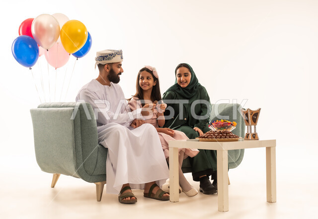 مفهوم الكرم وتقديم الضيافة العربية، طقوس الاحتفالات والمناسبات السعيدة في سلطنة عمان، جو البهجة والفرح والزينة، بورتريه لعائلة عربية خليجية عمانية تحتفل بالعيد المبارك، ارتداء الأزياء العمانية التقليدية، توزيع الحلويات الشعبية في الأعياد، خلفية بيضاء .