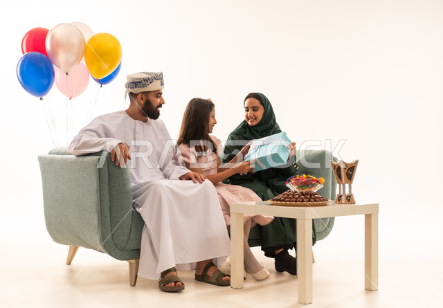 Gestures of satisfaction and pleasure, intimacy and affection between members of the Omani family, portrait of an Omani Gulf Arab couple sitting on the sofa with their baby girl, sharing and giving gifts on occasions and birthdays, family meetings and celebrations, white background.