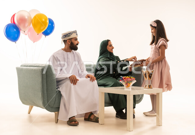 المحبة والمودة بين أفراد الأسرة، التهنئات وطقوس المناسبات الاسلامية، قيام الأبناء بتوزيع الضيافة، بنت عربية خليجية عمانية ترتدي ثياب العيد تقوم بتقديم الحلويات الشعبية، بورتريه لعائلة عربية خليجية عمانية تحتفل بقدوم العيد, الجلوس في غرفة المعيشة في جو عائلي، خلفية بيضاء .