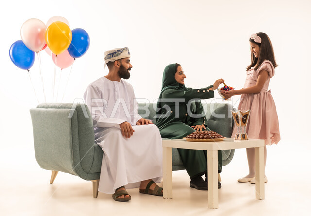 قيام الأبناء بتوزيع الضيافة، بنت عربية خليجية عمانية ترتدي ثياب العيد تقوم بتقديم الحلويات الشعبية، المحبة والمودة بين أفراد الأسرة، التهنئات وطقوس المناسبات الاسلامية، بورتريه لعائلة عربية خليجية عمانية تحتفل بقدوم العيد, الجلوس في غرفة المعيشة في جو عائلي، خلفية بيضاء .