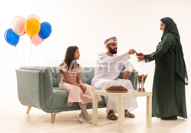 جو البهجة والفرح والزينة، طقوس الاحتفالات والمناسبات السعيدة في سلطنة عمان، توزيع الحلويات الشعبية في الأعياد، بورتريه لعائلة عربية خليجية عمانية تحتفل بالعيد المبارك، ارتداء الأزياء العمانية التقليدية، مفهوم الكرم وتقديم الضيافة العربية، خلفية بيضاء .