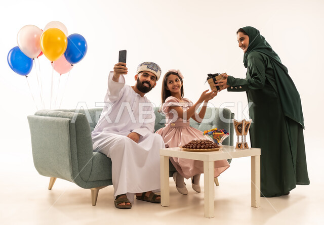 التقاط الصور التذكارية ، الألفة والمودة بين أفراد الأسرة العمانية ، بورتريه لزوجة عربية خليجية عمانية تقدم الهدية لطفلتها ، مشاركة وتقديم الهدايا بالمناسبات وأعياد الميلاد, ايماءات الرضا والسرور، الاجتماعات والاحتفالات العائلية، خلفية بيضاء .