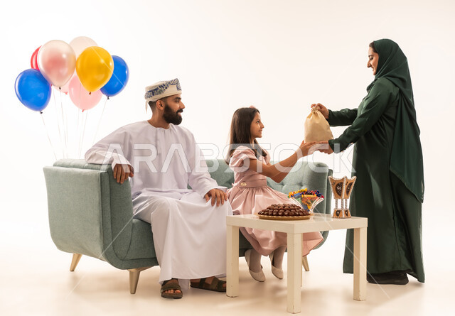 تجمع العائلة العمانية لاستقبال العيد المبارك, إظهار مشاعر الحنان والمحبة, الجلوس في غرفة المعيشة في جو عائلي دافئ، بورتريه لعائلة عربية خليجية عمانية تحتفل بالمناسبات الاسلامية برفقة بعضها البعض, ارتداء الثياب الجديدة الجميلة ، خلفية بيضاء .