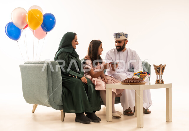 جو البهجة والفرح والزينة، الزيارات العائلية وتجمعات الأسر ، تقديم العيديات و الحلوى ، تقديم العيدية للأطفال في عيد الفطر المبارك ، بورتريه لعائلة عمانية داخل المنزل تحتفل بعيد الفطر السعيد ، عيد فطر مبارك ، خلفية بيضاء .