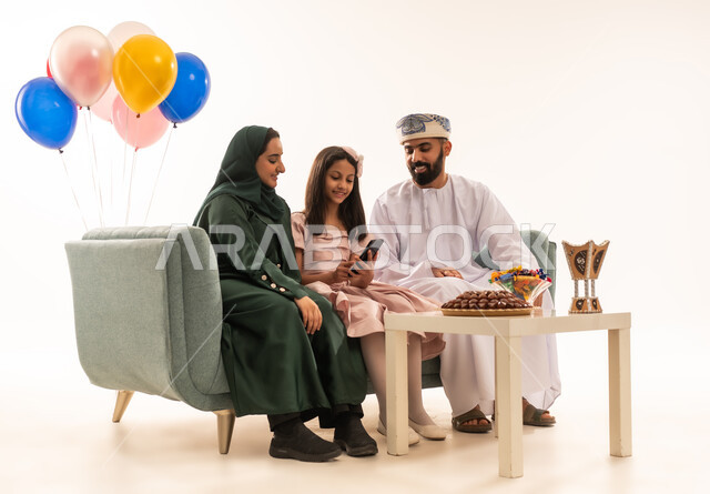 تصفح مواقع التواصل الاجتماعى باستخدام الجوال، الجلوس في غرفة المعيشة في جو عائلي دافئ، تجمع العائلة العمانية لاستقبال العيد المبارك, إظهار مشاعر الحنان والمحبة,  بورتريه لعائلة عربية خليجية عمانية تحتفل بالمناسبات الاسلامية برفقة بعضها البعض, ارتداء الثياب الجديدة الجميلة ، خلفية بيضاء .