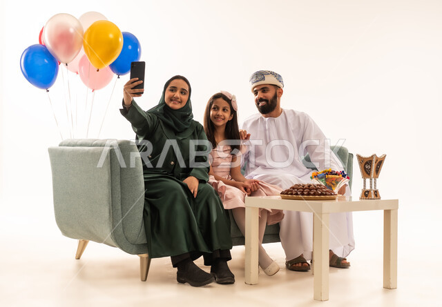 اجراء مكالمات الفيديو فى العيد، الجلوس في غرفة المعيشة في جو عائلي دافئ، تجمع العائلة العمانية لاستقبال العيد المبارك, إظهار مشاعر الحنان والمحبة,  بورتريه لعائلة عربية خليجية عمانية تحتفل بالمناسبات الاسلامية برفقة بعضها البعض, ارتداء الثياب الجديدة الجميلة ، خلفية بيضاء .