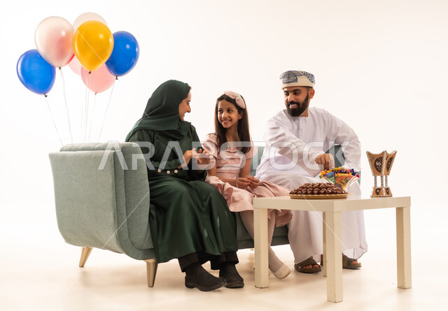 ضيافات الأعياد العربية الشعبية في سلطنة عمان، الاحتفال بالأعياد الإسلامية والزيارات العائلية، بورتريه لعائلة عربية خليجية عمانية تجلس في غرفة المعيشة وتقوم بتبادل المعايدات، إظهار مشاعر الفرح والسعادة بقدوم العيد، مفهوم الحنان والمحبة والمودة، خلفية بيضاء .