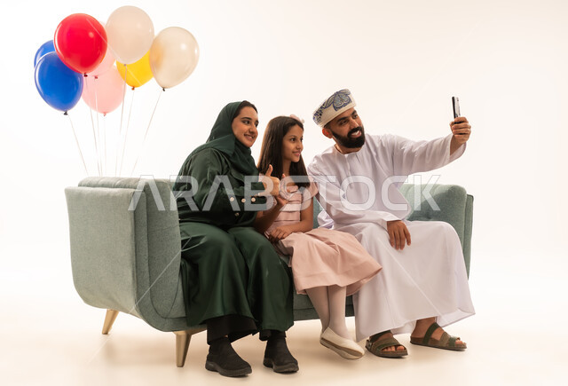 النظر الى كاميرا السيلفي بايماءات السعادة، الجلوس في غرفة المعيشة في جو عائلي دافئ، تجمع العائلة العمانية لاستقبال العيد المبارك, إظهار مشاعر الحنان والمحبة,  بورتريه لعائلة عربية خليجية عمانية تحتفل بالمناسبات الاسلامية برفقة بعضها البعض, ارتداء الثياب الجديدة الجميلة ، خلفية بيضاء .