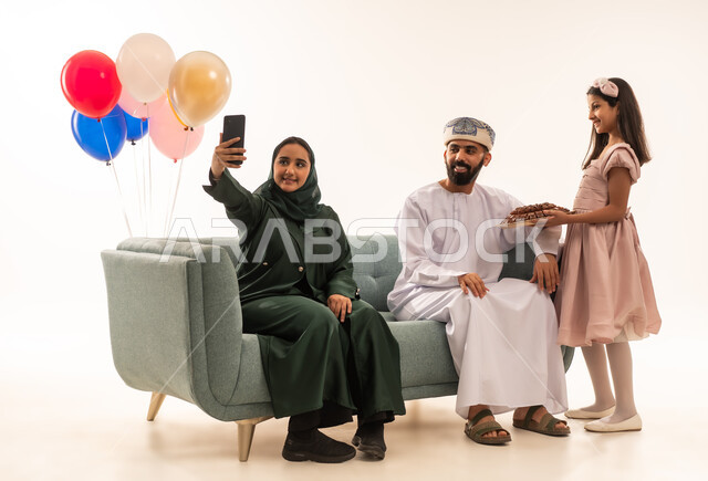 طقوس الاحتفالات والمناسبات السعيدة, الاحتفال بالأعياد الإسلامية والزيارات العائلية، بنت عربية خليجية عمانية ترتدي ثياب العيد تقوم بتقديم الحلويات الشعبية، صور السيلفي التذكارية ، بورتريه لعائلة عربية خليجية عمانية تجلس في غرفة المعيشة وتقوم بتبادل المعايدات، مفهوم الحنان والمحبة والمودة، خلفية بيضاء .